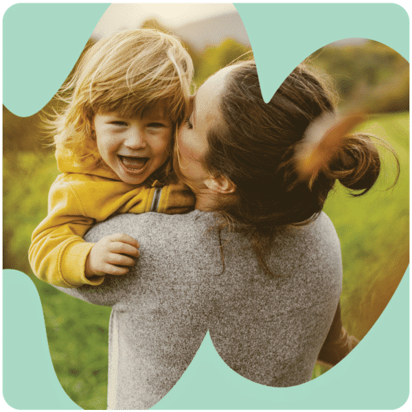 A woman in a gray sweater kisses a laughing toddler in a yellow jacket outdoors, framed by a heart-shaped cutout.