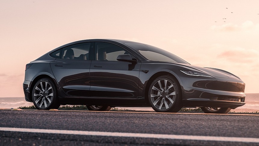 A sleek black electric car is parked on a road, with the ocean and a pastel-colored sky in the background.