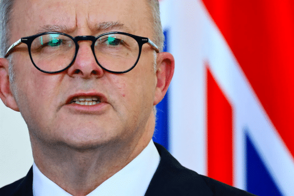A man wearing glasses with a national flag in the background.
