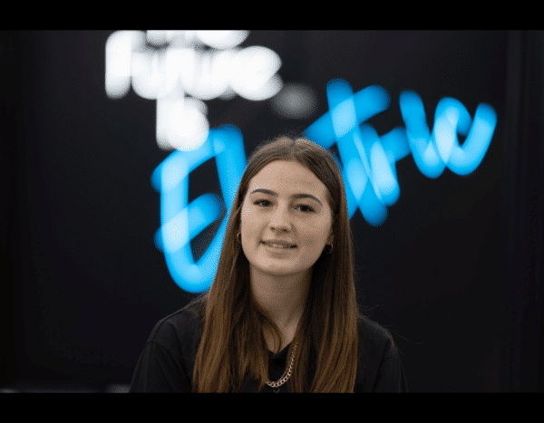 Young woman with a neutral expression standing in front of a black background with the illuminated words "the future is bright.